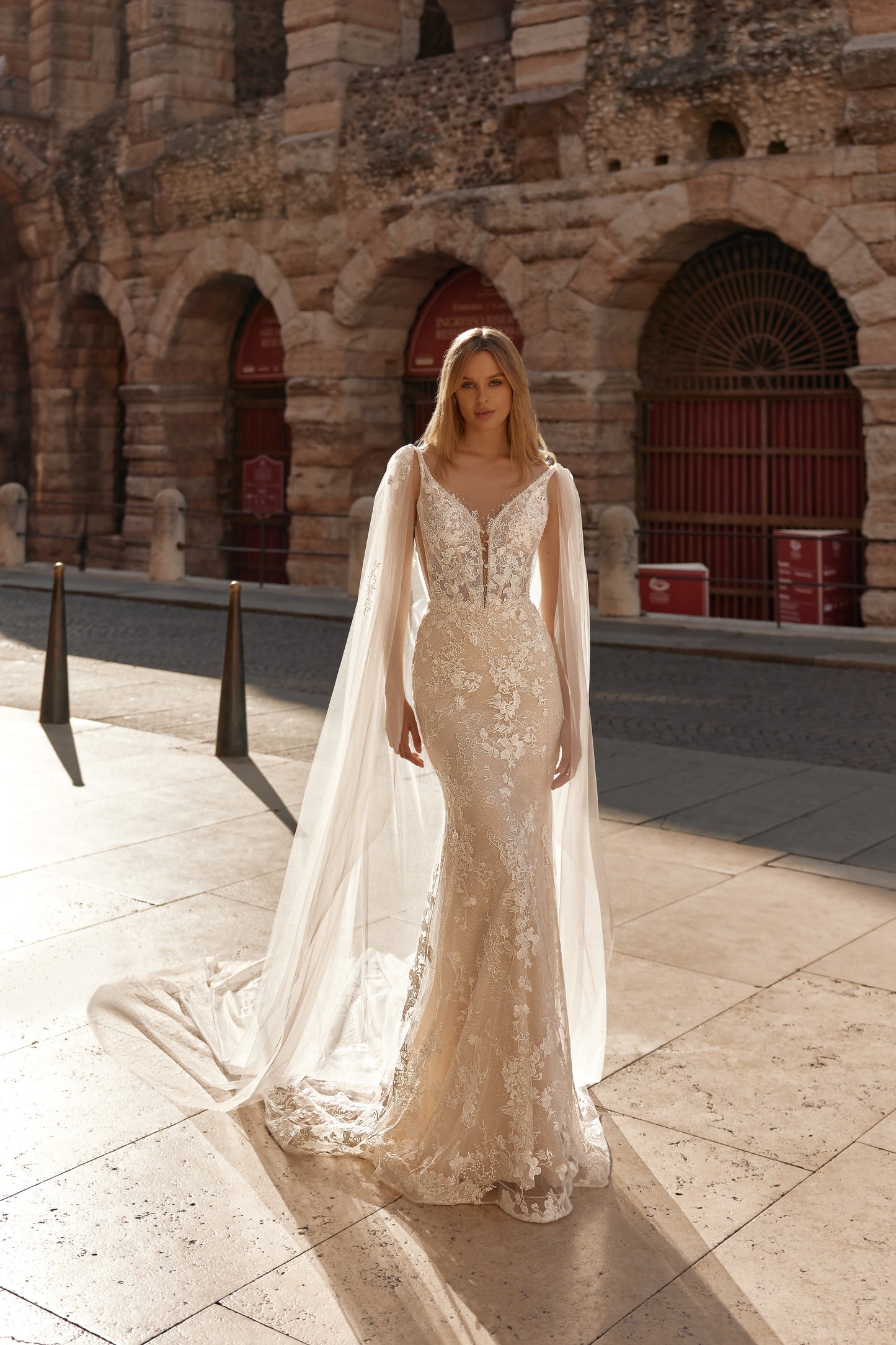 Woman in a white lace gown with a cape standing in front of an ancient stone building.