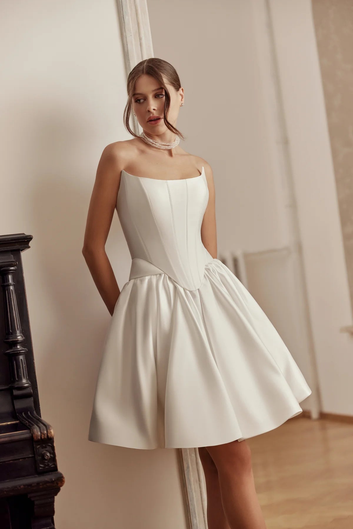 Bride in elegant tea-length satin gown with a square neckline and flared skirt, standing in a softly lit room.