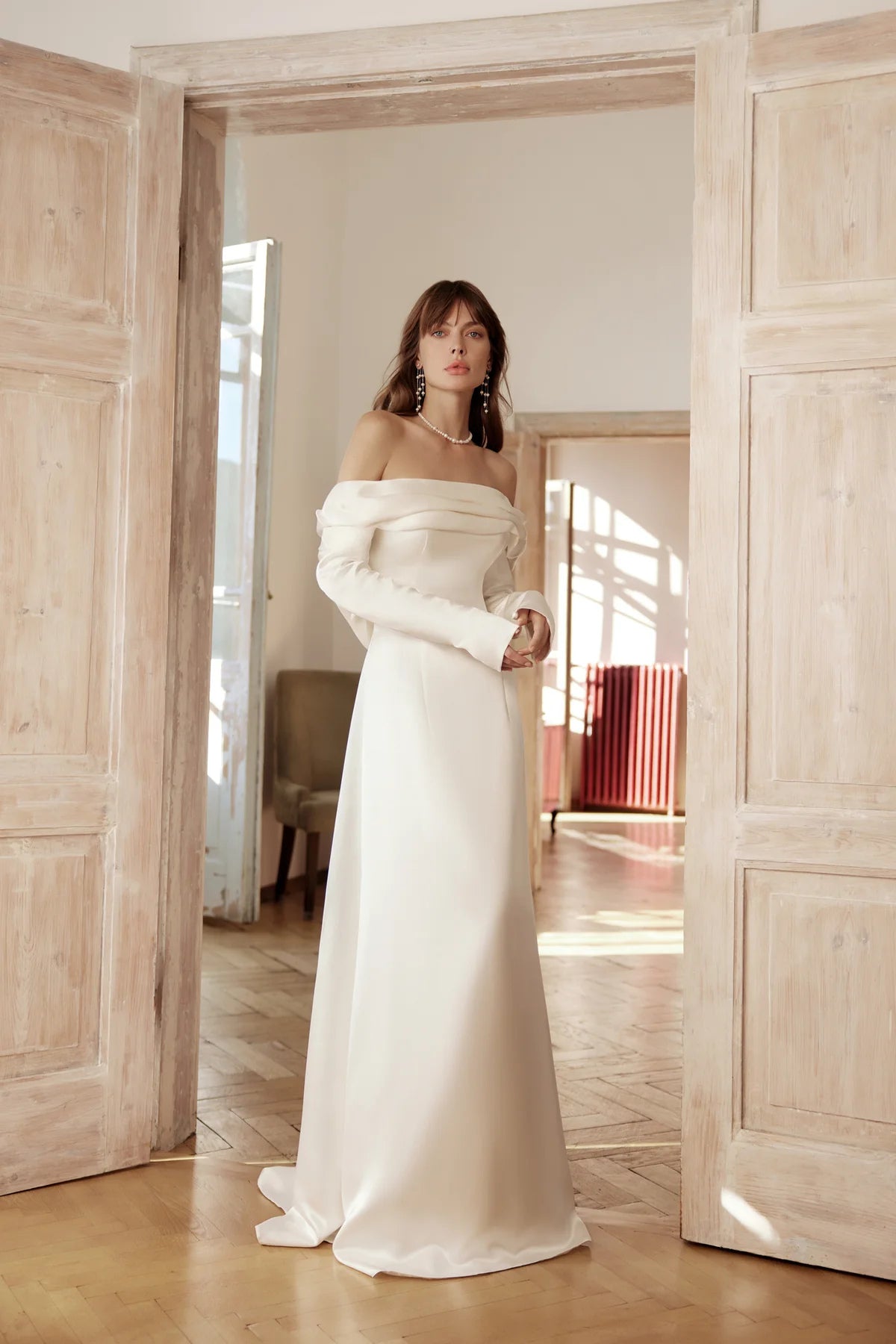 Bride in ivory off-the-shoulder silk gown with draped sleeves, standing in soft light