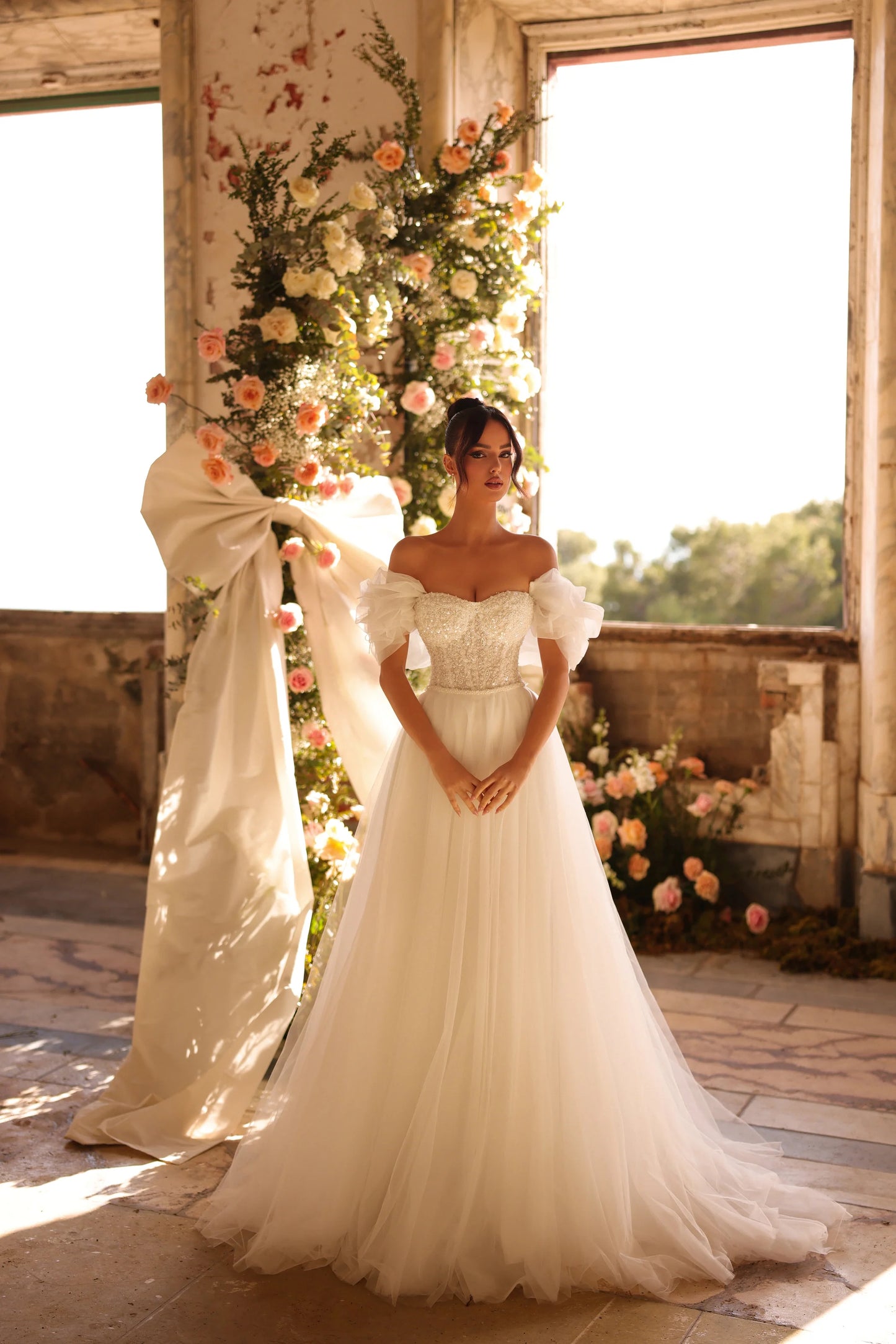 Bride in sparkling gown with airy tulle skirt and puffed sleeves near floral arch