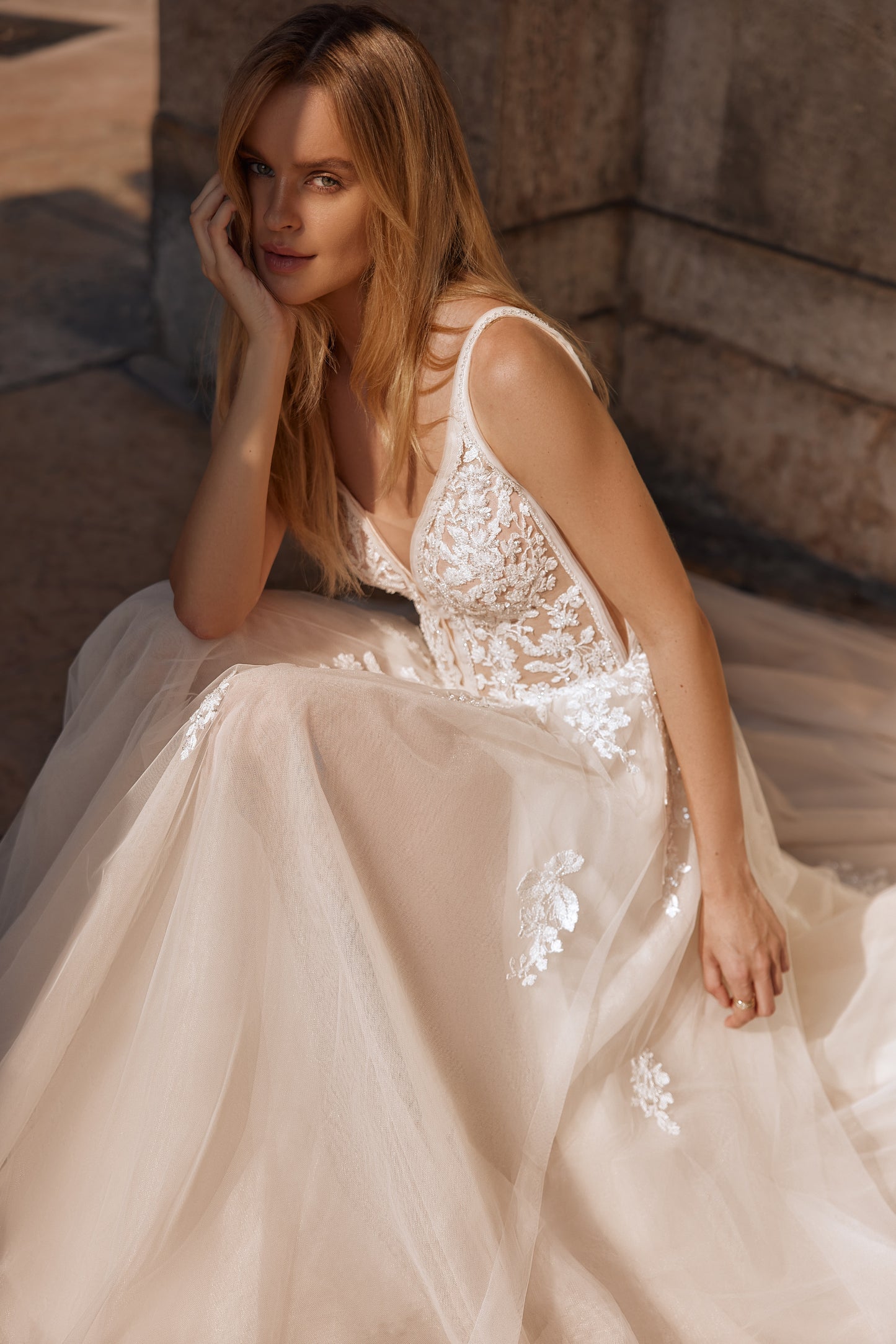 Woman in a beige wedding dress with floral details sitting on a stone surface.