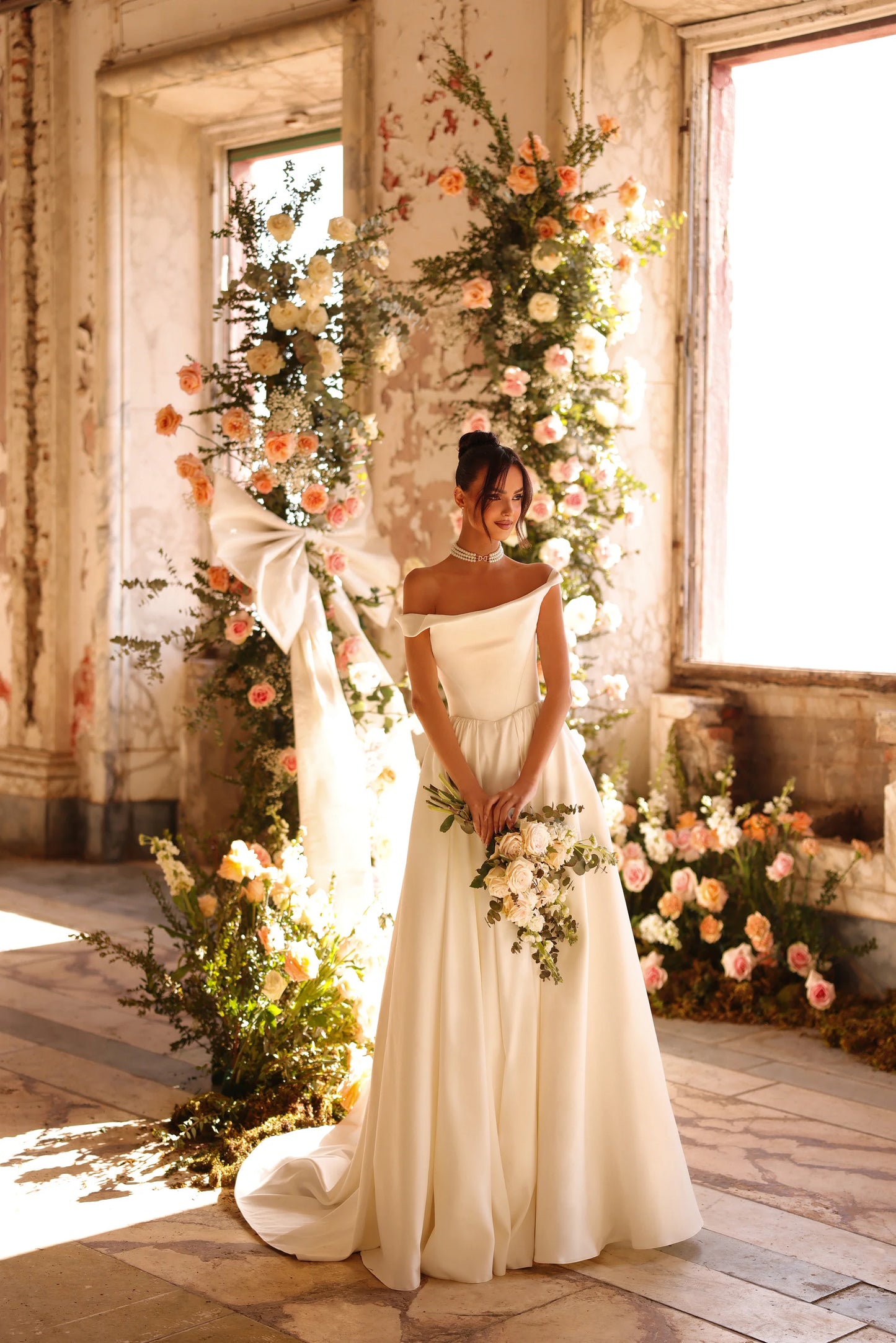 Bride in satin wedding gown with off-the-shoulder neckline standing near window