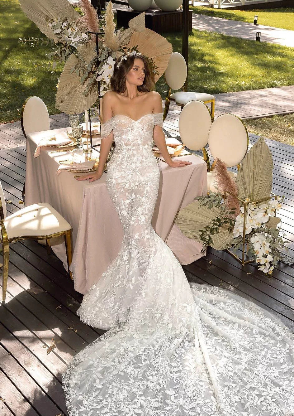Bride wearing a semi-transparent mermaid gown with lace details, posing with a chic hat and bouquet in a garden setting.