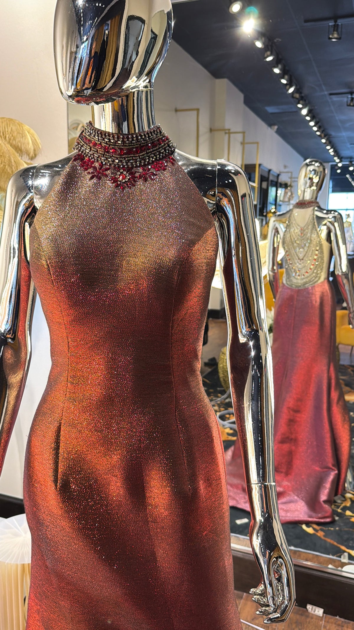 A metallic bronze evening gown with a beaded red and black collar, displayed on a silver mannequin in a boutique showroom.