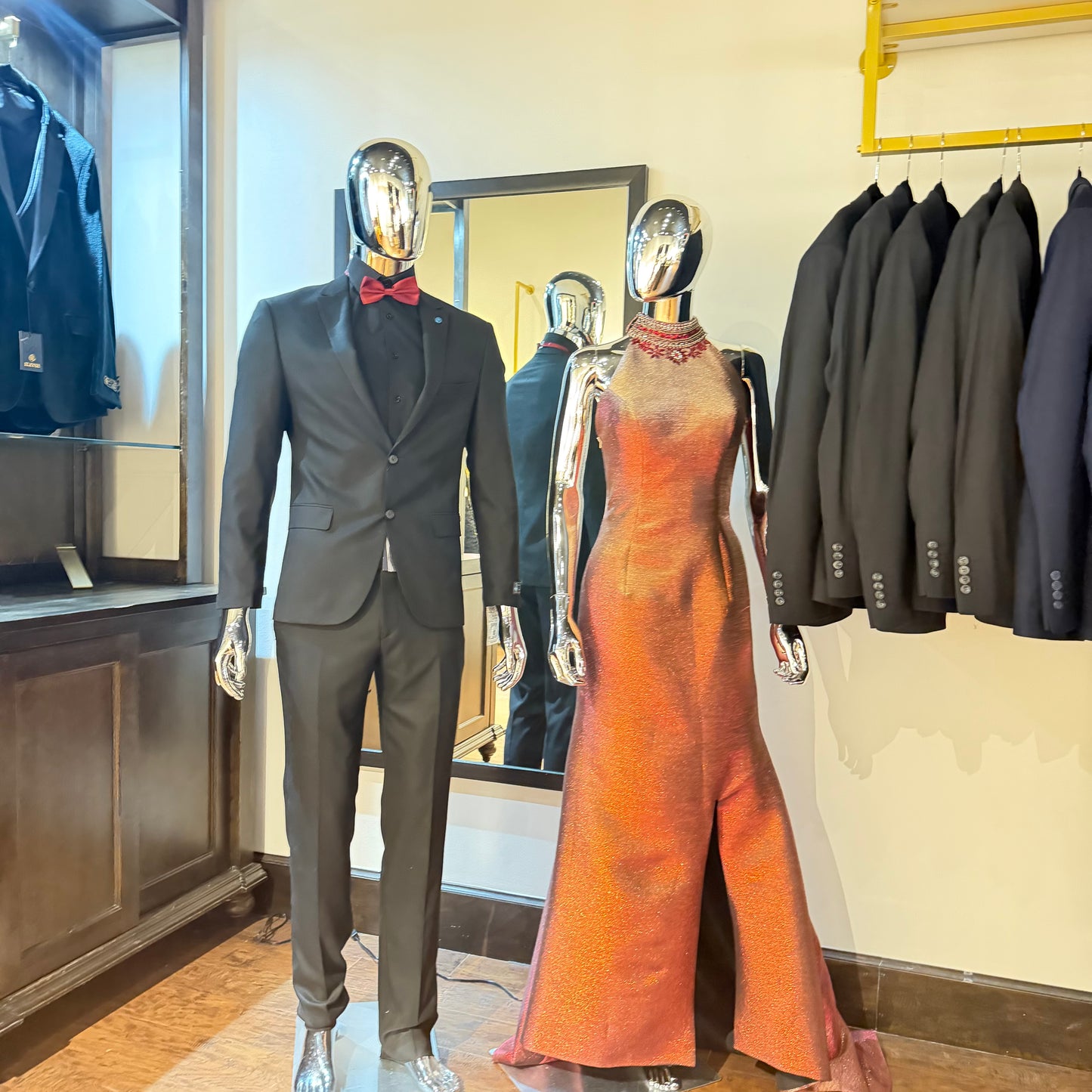A mannequin wearing a dark gray suit with a red bow tie stands beside a metallic mannequin in a sleeveless orange-red evening gown inside a clothing showroom. Formalwear garments are hanging on a rack in the background.