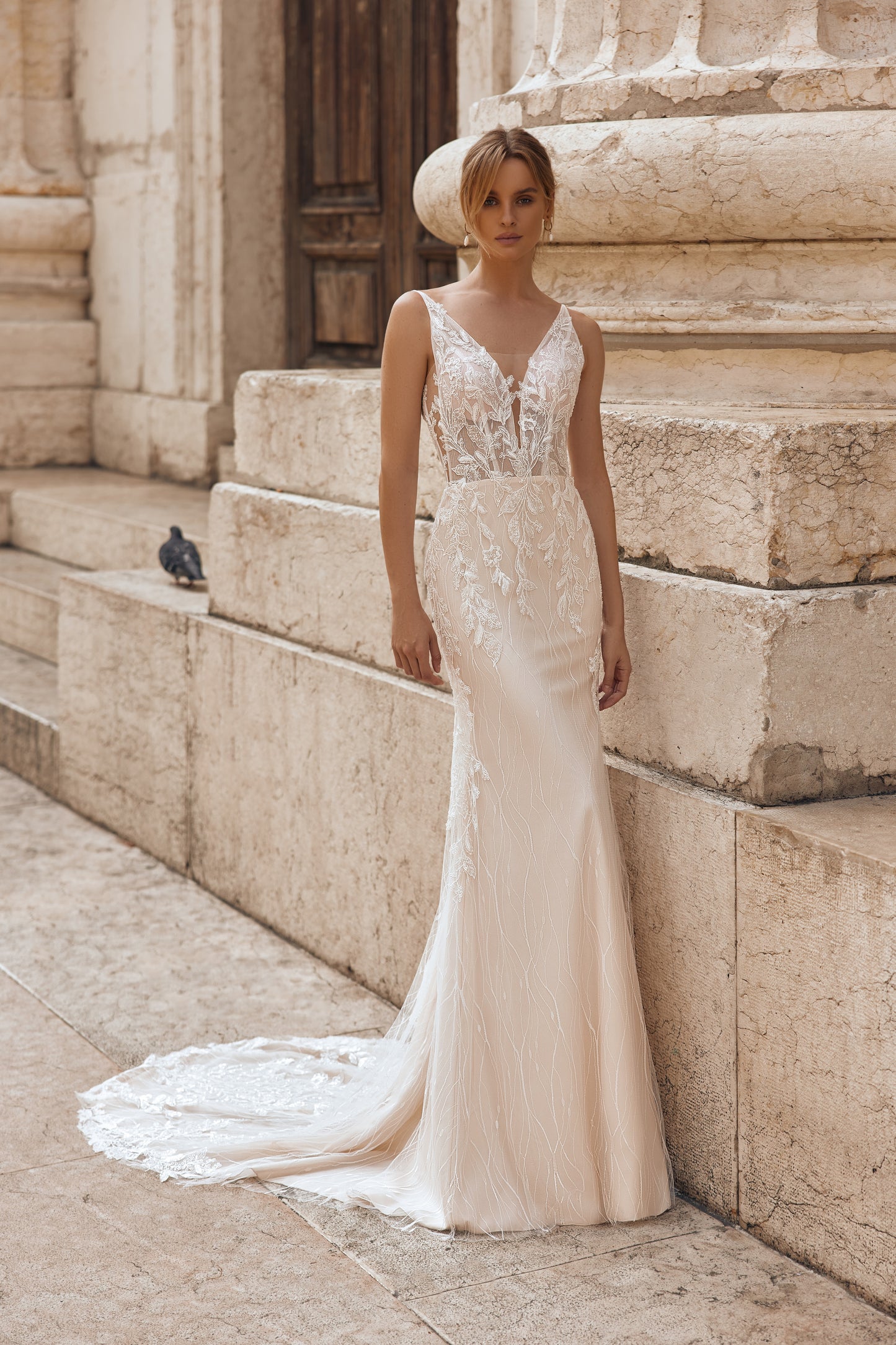 Woman in a white wedding dress standing on stone steps with a pigeon nearby.