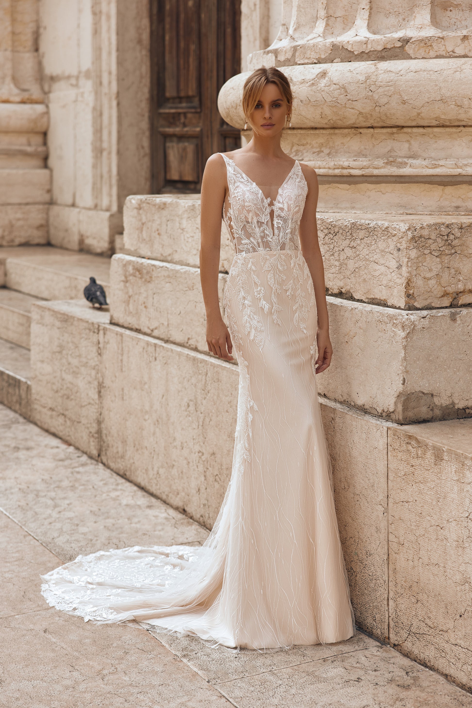 Woman in a white wedding dress standing on stone steps with a pigeon nearby.