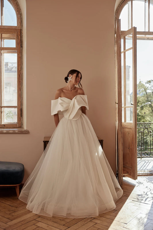 Bride in off-shoulder ball gown with full tulle skirt, posing near window