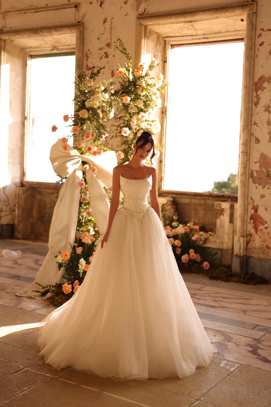 Bride in a romantic ball gown standing by a large window, soft natural light illuminating the tulle skirt.