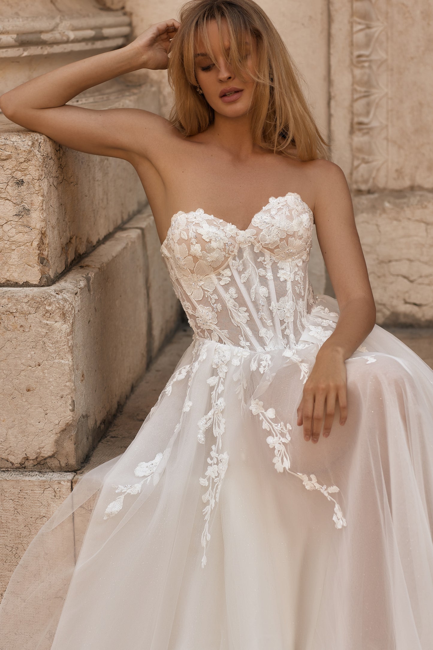 Woman in a strapless wedding dress with floral details against a stone wall.