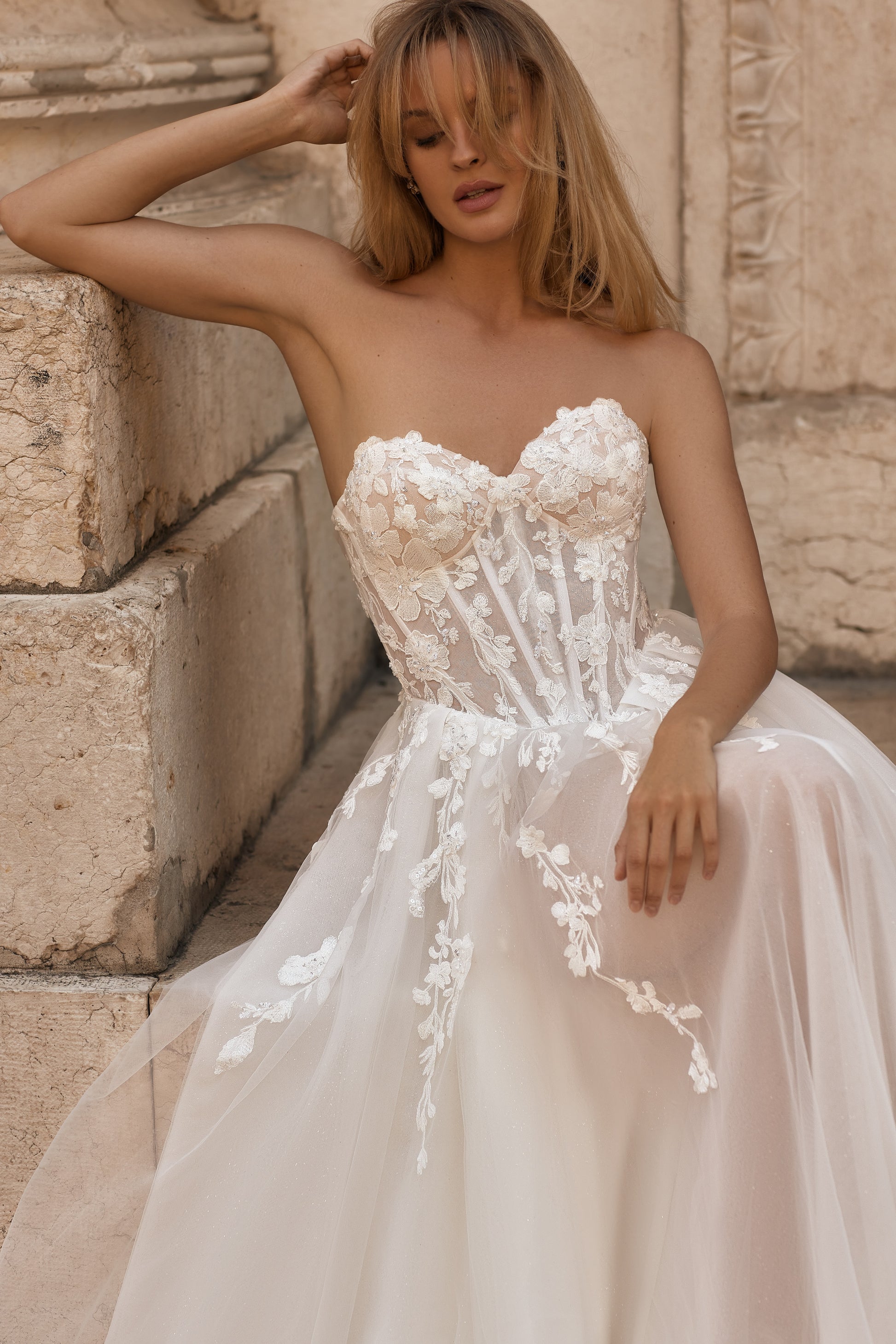 Woman in a strapless wedding dress with floral details against a stone wall.