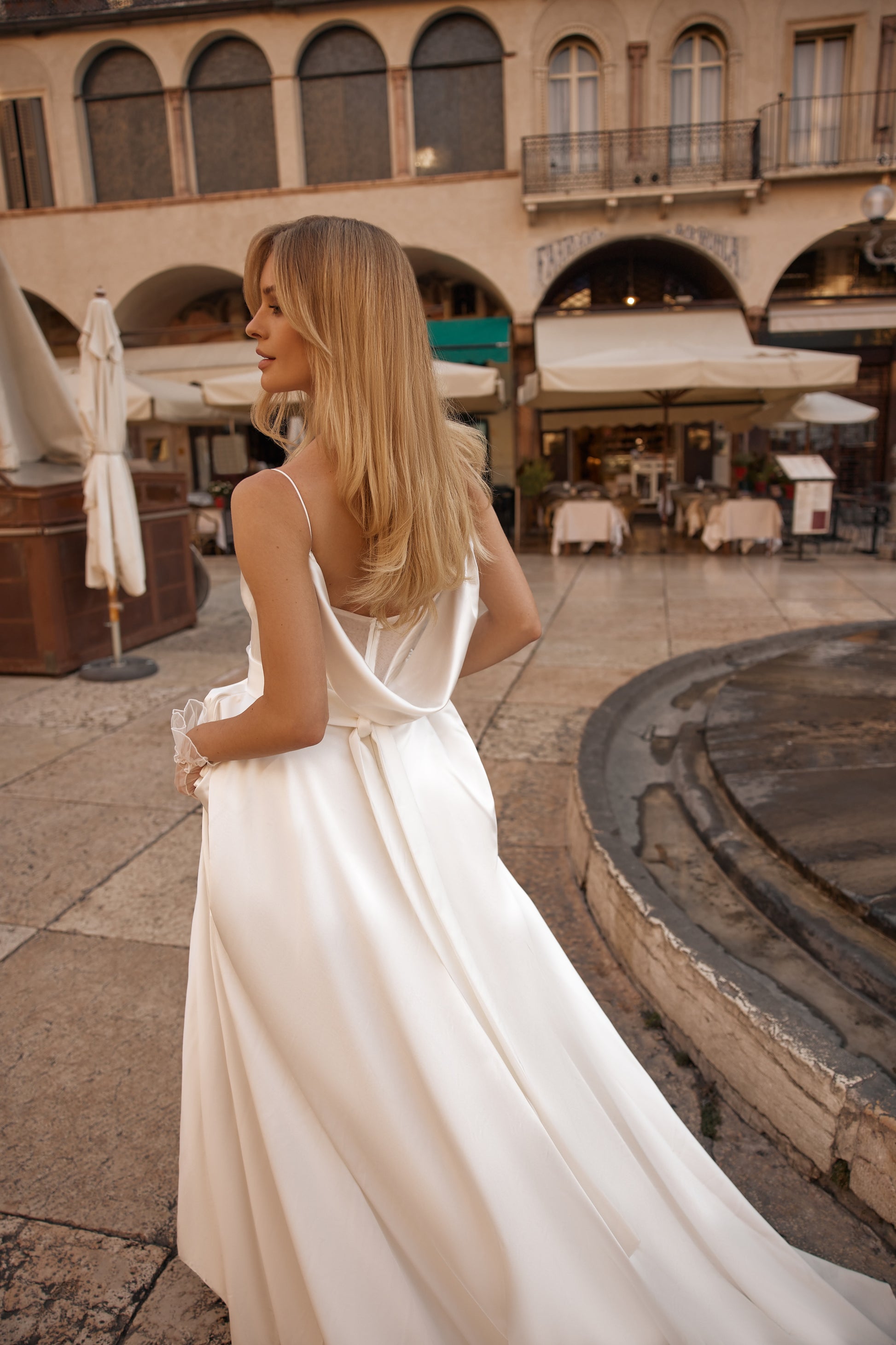 Woman in a white dress standing in an outdoor setting with arches and tables.