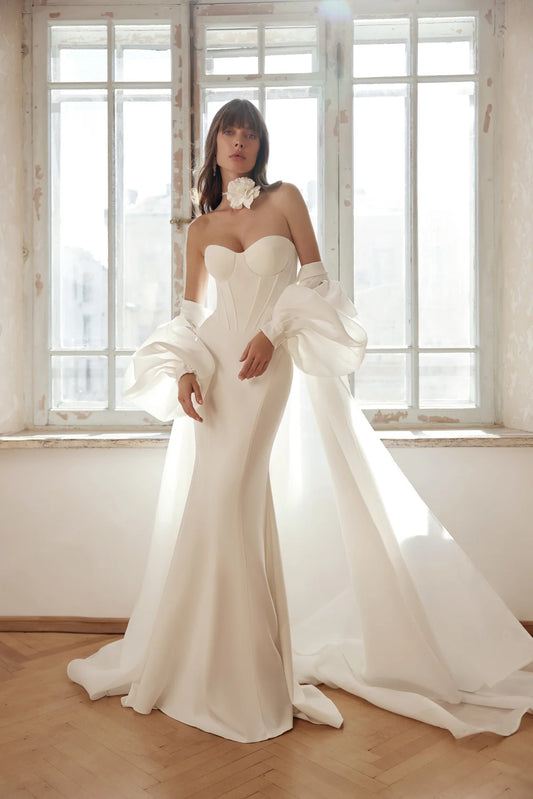 A stunning bride in an ivory mermaid wedding gown with a corset bodice, dramatic train, and detachable puffed sleeves, standing near a window in soft natural light.