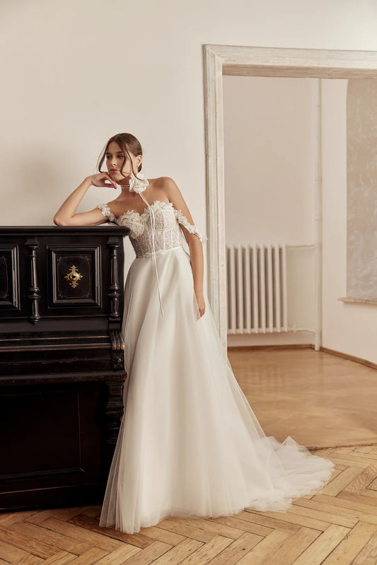 Bride wearing an ivory A-line gown with floral lace and off-shoulder sleeves, standing by a classic piano.