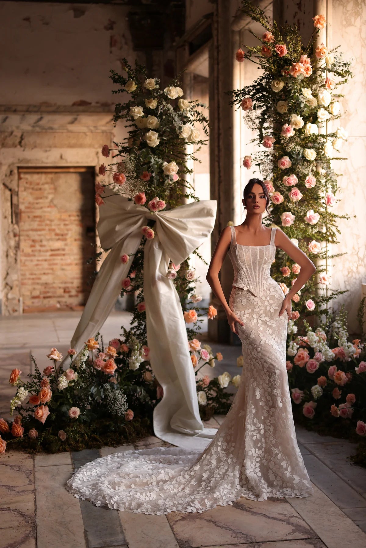 Bride standing in front of floral arch in a fitted wedding dress with floral lace details, hands on hips, looking confident