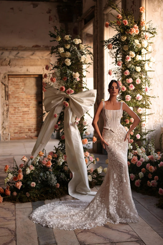 Bride standing in front of floral arch in a fitted wedding dress with floral lace details, hands on hips, looking confident