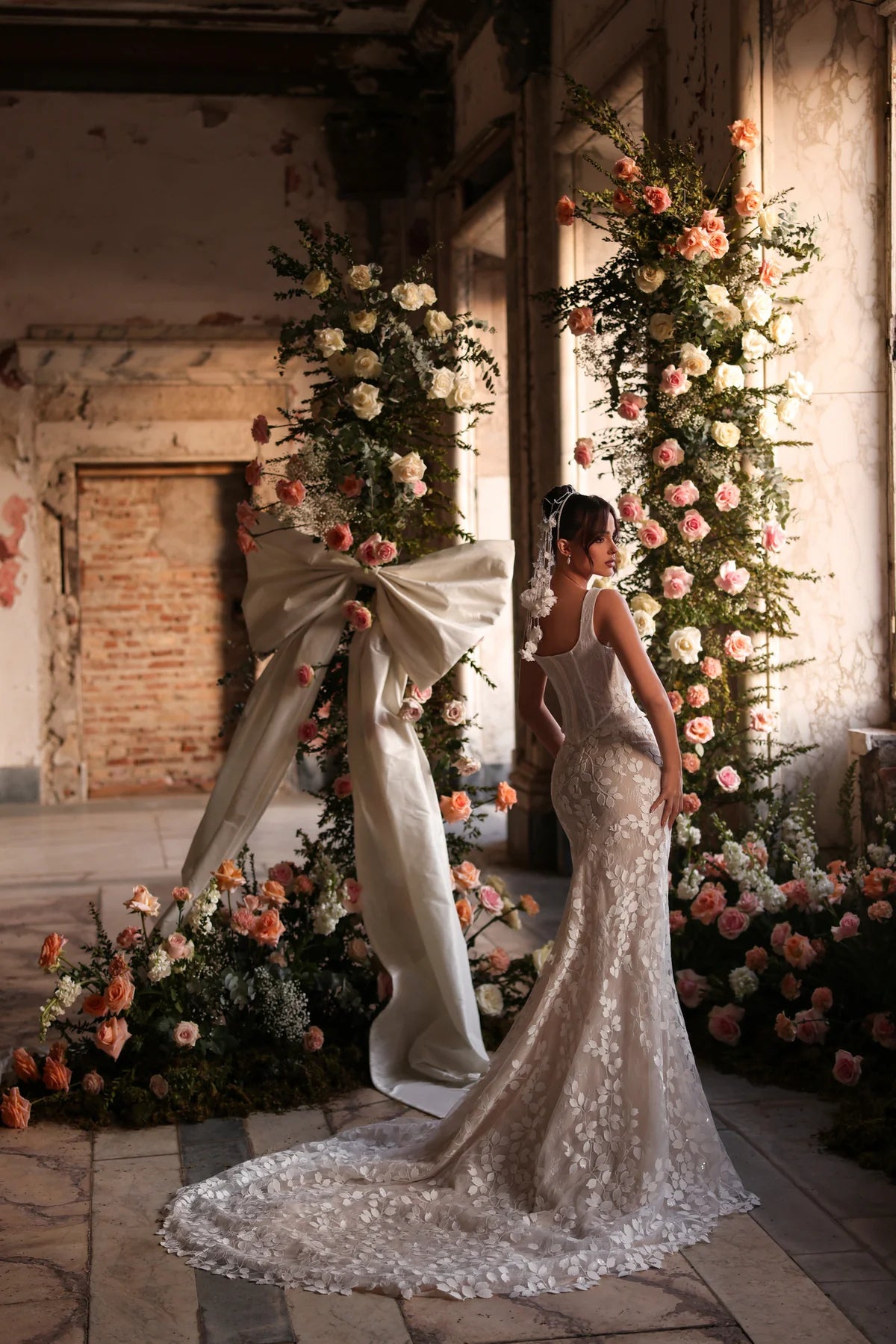 Bride in a floral lace wedding dress, turned slightly to show the back, soft train trailing behind, surrounded by pastel flower arrangements