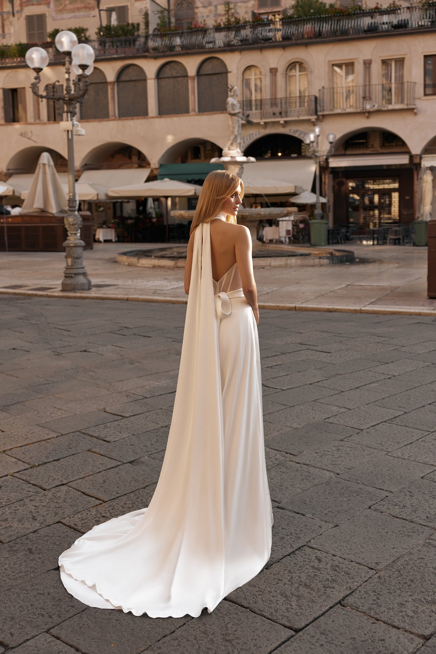Woman in a white dress with a long train standing on a stone pavement in an urban setting.