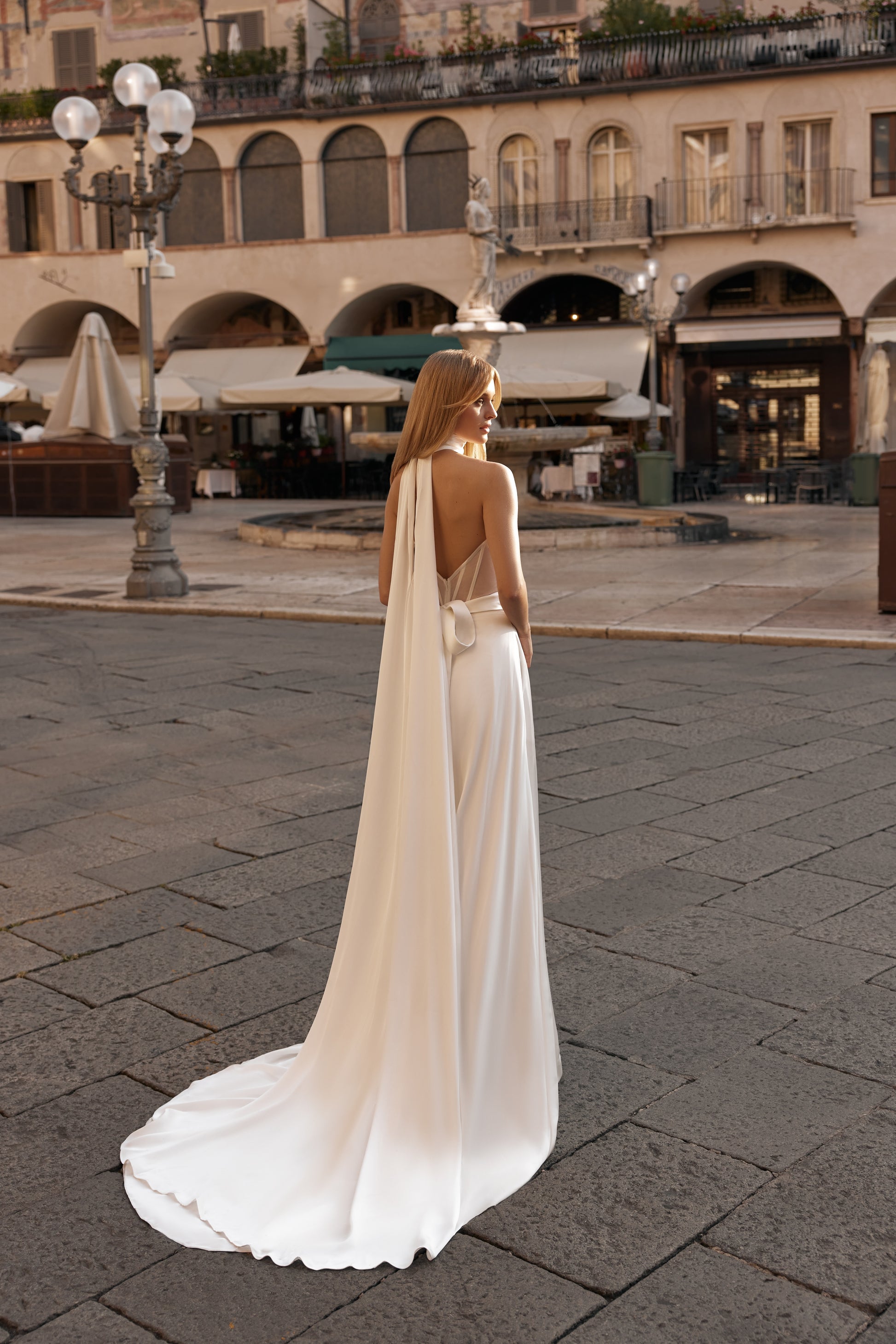 Woman in a white dress with a long train standing on a stone pavement in an urban setting.